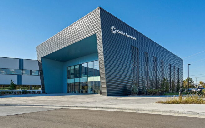 A modern, angular building with dark metal paneling and large glass windows. The sign reads “Collins Aerospace.” A wide sidewalk, greenery, and a clear blue sky are visible.