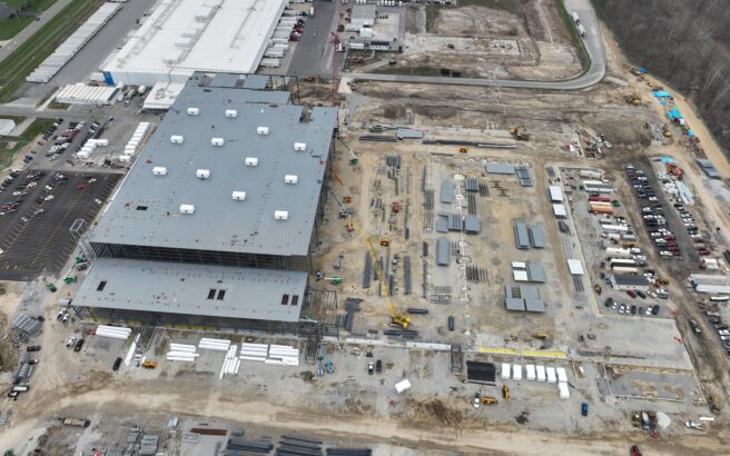 Aerial view of an industrial construction site with a large building, scattered materials, vehicles, and equipment, surrounded by parking lots and partially developed land. Trees border the site on one side.