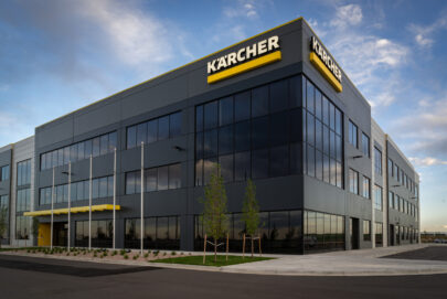 A modern, three-story office building with large glass windows and a yellow Kärcher sign on the corner. The area is landscaped with small trees and the sky is partly cloudy.