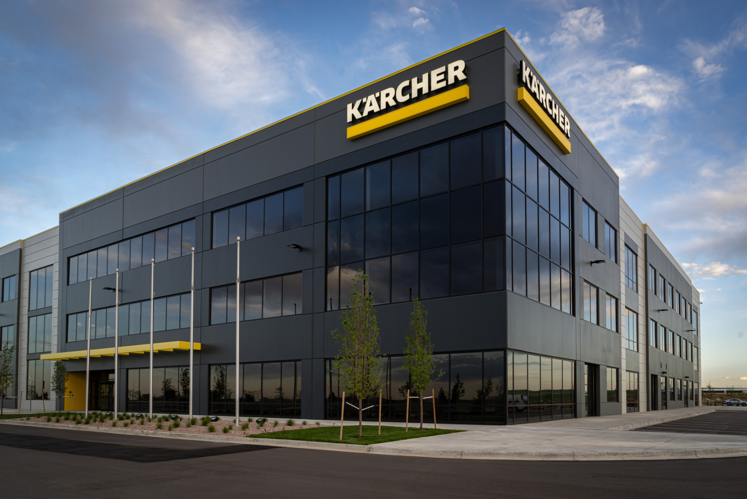 A modern, three-story office building with large glass windows and a yellow Kärcher sign on the corner. The area is landscaped with small trees and the sky is partly cloudy.