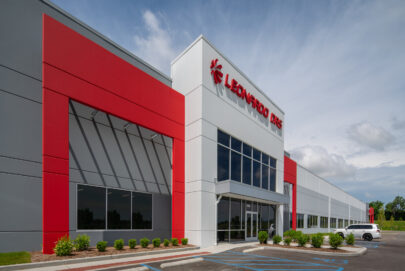 Modern commercial building with a red and white facade, large glass windows, and Leonardo DRS signage above the entrance. There are landscaped bushes in front and a white car parked nearby under a partly cloudy sky.