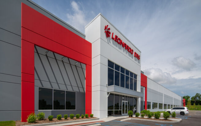 Modern commercial building with a red and white facade, large glass windows, and Leonardo DRS signage above the entrance. There are landscaped bushes in front and a white car parked nearby under a partly cloudy sky.