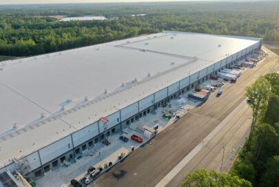A large industrial warehouse with multiple loading docks is surrounded by trees and vehicles, viewed from above on a sunny day.