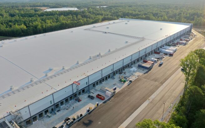A large industrial warehouse with multiple loading docks is surrounded by trees and vehicles, viewed from above on a sunny day.