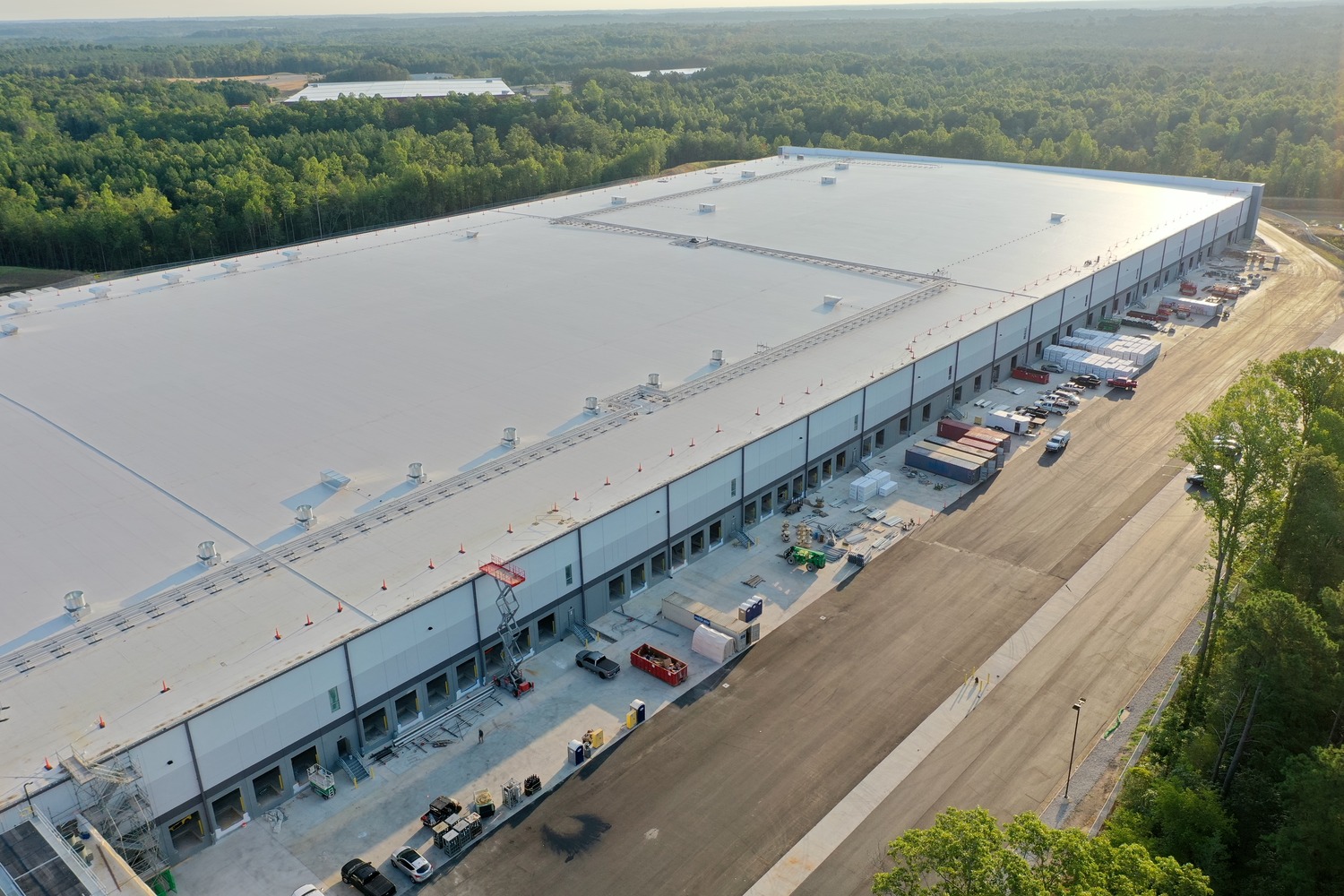 A large industrial warehouse with multiple loading docks is surrounded by trees and vehicles, viewed from above on a sunny day.