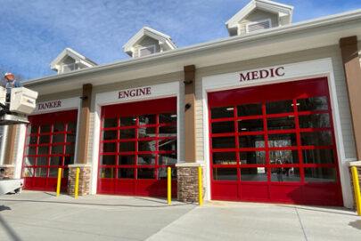 A modern fire station with three red garage doors labeled TANKER, ENGINE, and MEDIC under gabled roof peaks, set against a blue sky.