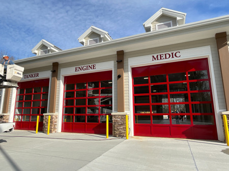 A modern fire station with three red garage doors labeled TANKER, ENGINE, and MEDIC under gabled roof peaks, set against a blue sky.