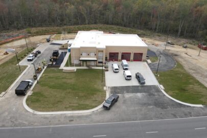 Aerial view of a modern fire station with three red garage doors, several parked vehicles, and workers paving the driveway. The building is surrounded by grass and bordered by a forested area in the background.