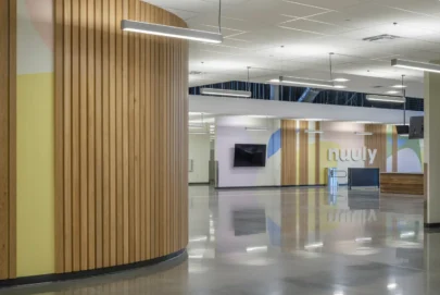 Modern office interior with curved wooden wall panels, polished concrete floors, suspended linear lights, and pastel-colored wall art. A TV is mounted on the wall, and the word nuuly is displayed in large letters.