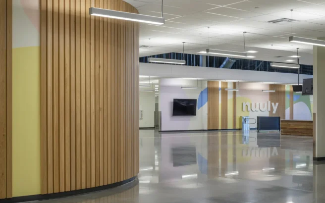 Modern office interior with curved wooden wall panels, polished concrete floors, suspended linear lights, and pastel-colored wall art. A TV is mounted on the wall, and the word nuuly is displayed in large letters.