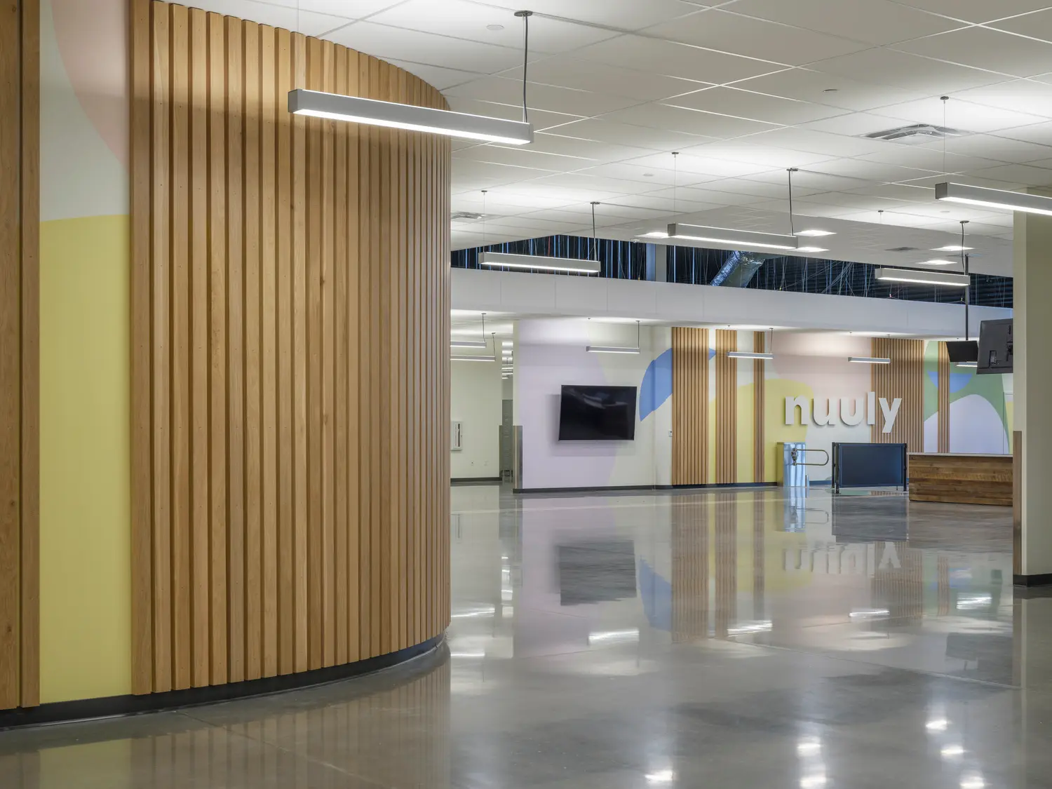 Modern office interior with curved wooden wall panels, polished concrete floors, suspended linear lights, and pastel-colored wall art. A TV is mounted on the wall, and the word nuuly is displayed in large letters.