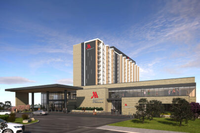 A modern Marriott hotel building with large glass windows, a tall main tower, and the red Marriott logo on the exterior, set against a blue sky with landscaped greenery and parked cars in front.