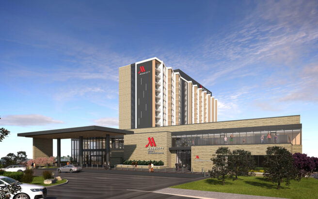 A modern Marriott hotel building with large glass windows, a tall main tower, and the red Marriott logo on the exterior, set against a blue sky with landscaped greenery and parked cars in front.