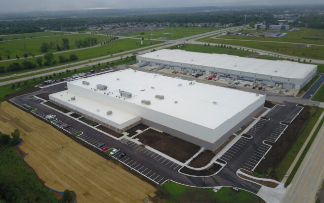Aerial view of a large, modern warehouse or distribution center with white roofs, surrounded by parking lots, loading docks, green fields, roads, and a nearby residential area.