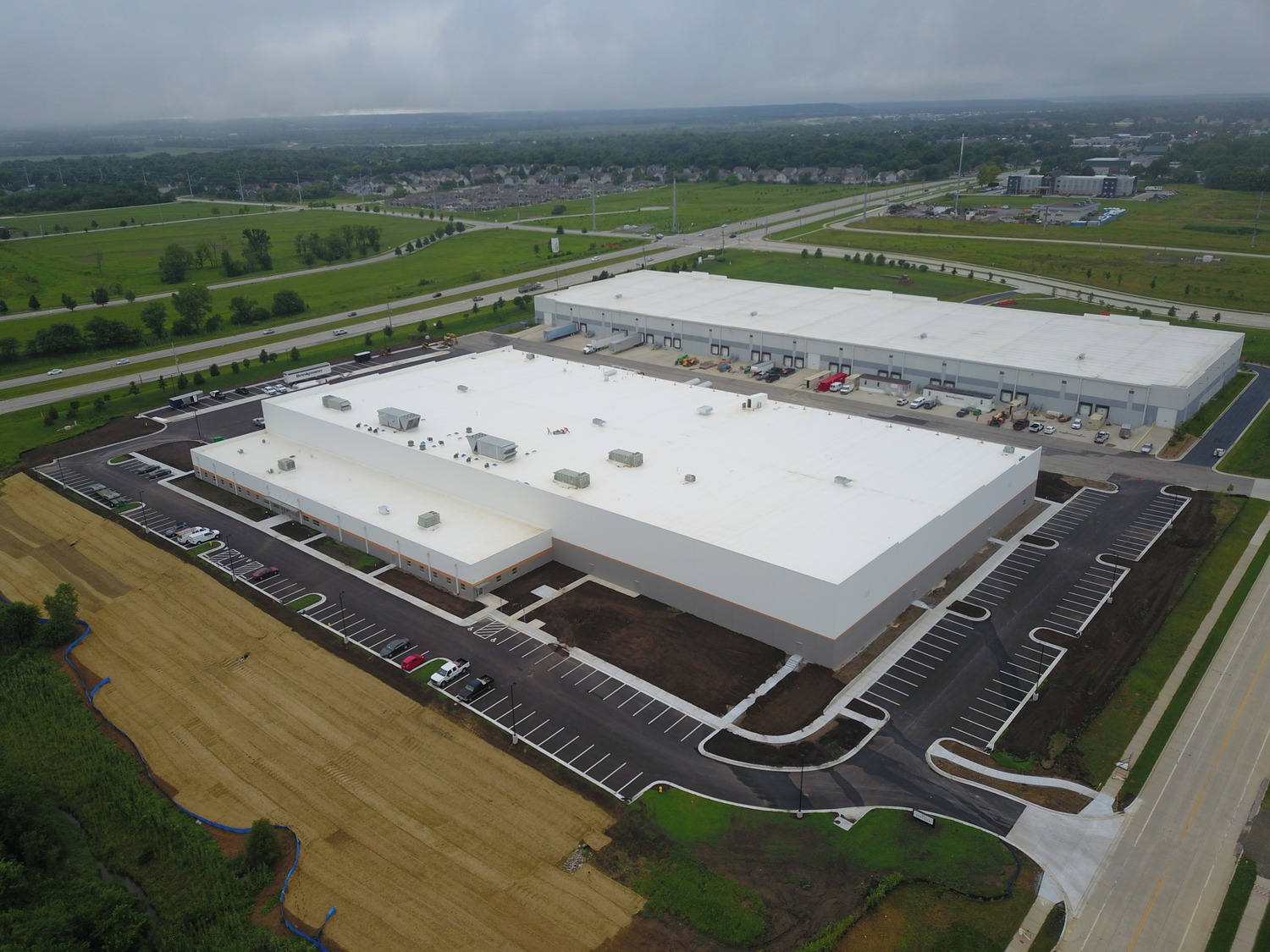 Aerial view of a large, modern warehouse or distribution center with white roofs, surrounded by parking lots, loading docks, green fields, roads, and a nearby residential area.
