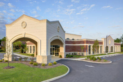 A modern, single-story building with beige and red brick exterior, arched entrance, large windows, neatly landscaped grounds, and an empty parking lot under a partly cloudy sky.