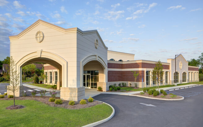 A modern, single-story building with beige and red brick exterior, arched entrance, large windows, neatly landscaped grounds, and an empty parking lot under a partly cloudy sky.