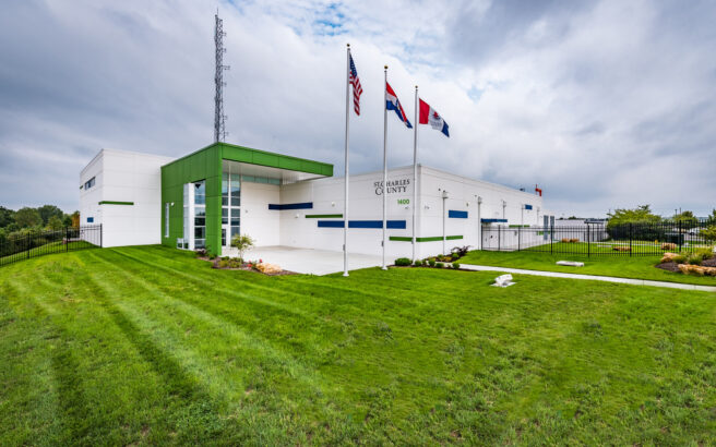 A modern white and green building with three flagpoles outside, displaying the U.S., Missouri, and another flag. The sign reads St. Charles County. The building is surrounded by a well-kept lawn and a cloudy sky above.