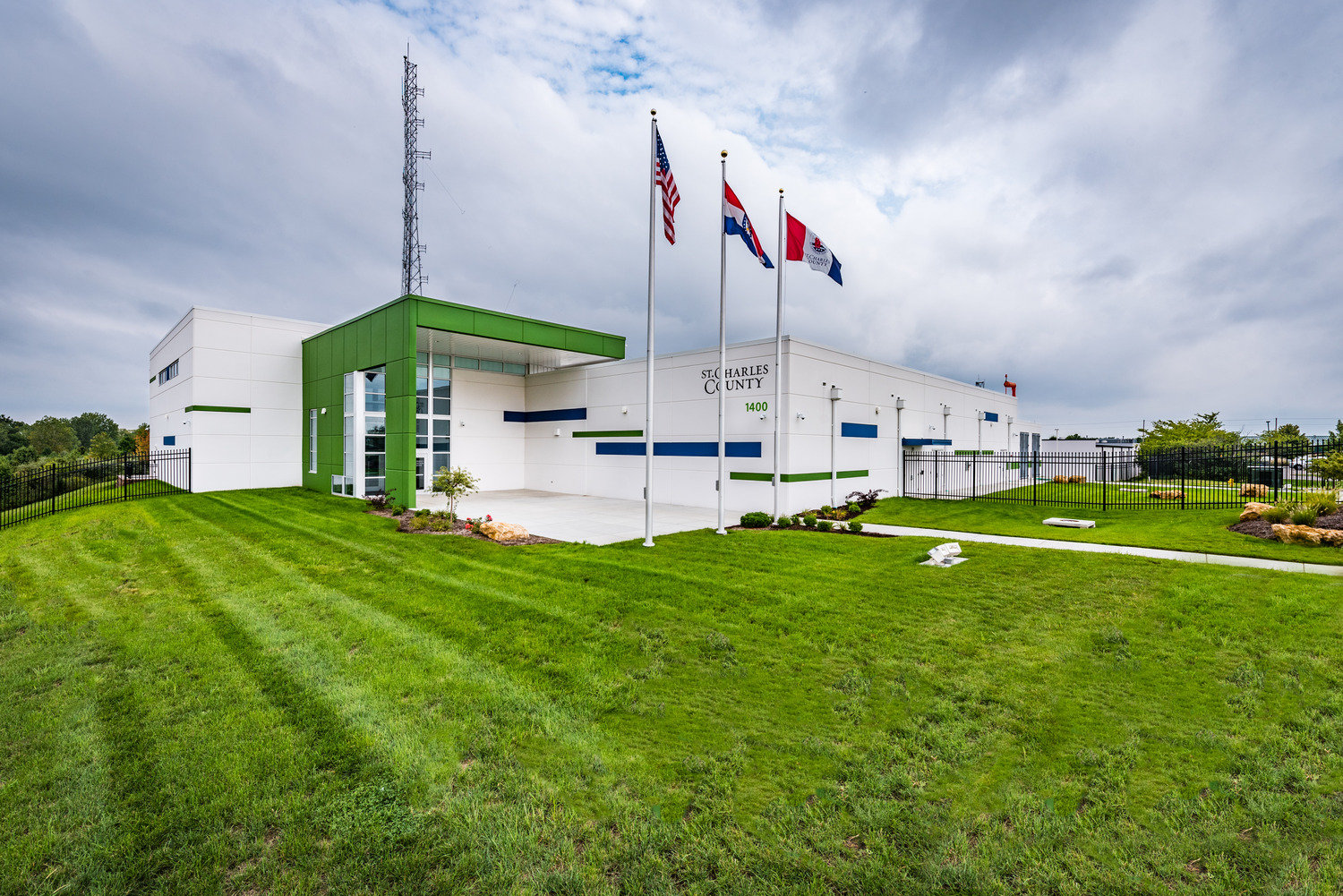 A modern white and green building with three flagpoles outside, displaying the U.S., Missouri, and another flag. The sign reads St. Charles County. The building is surrounded by a well-kept lawn and a cloudy sky above.