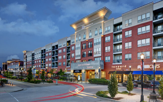 Modern mixed-use building with retail stores and restaurants on the ground floor and multiple stories of apartments above, photographed at dusk with streetlights, trees, and light trails from passing cars in front.