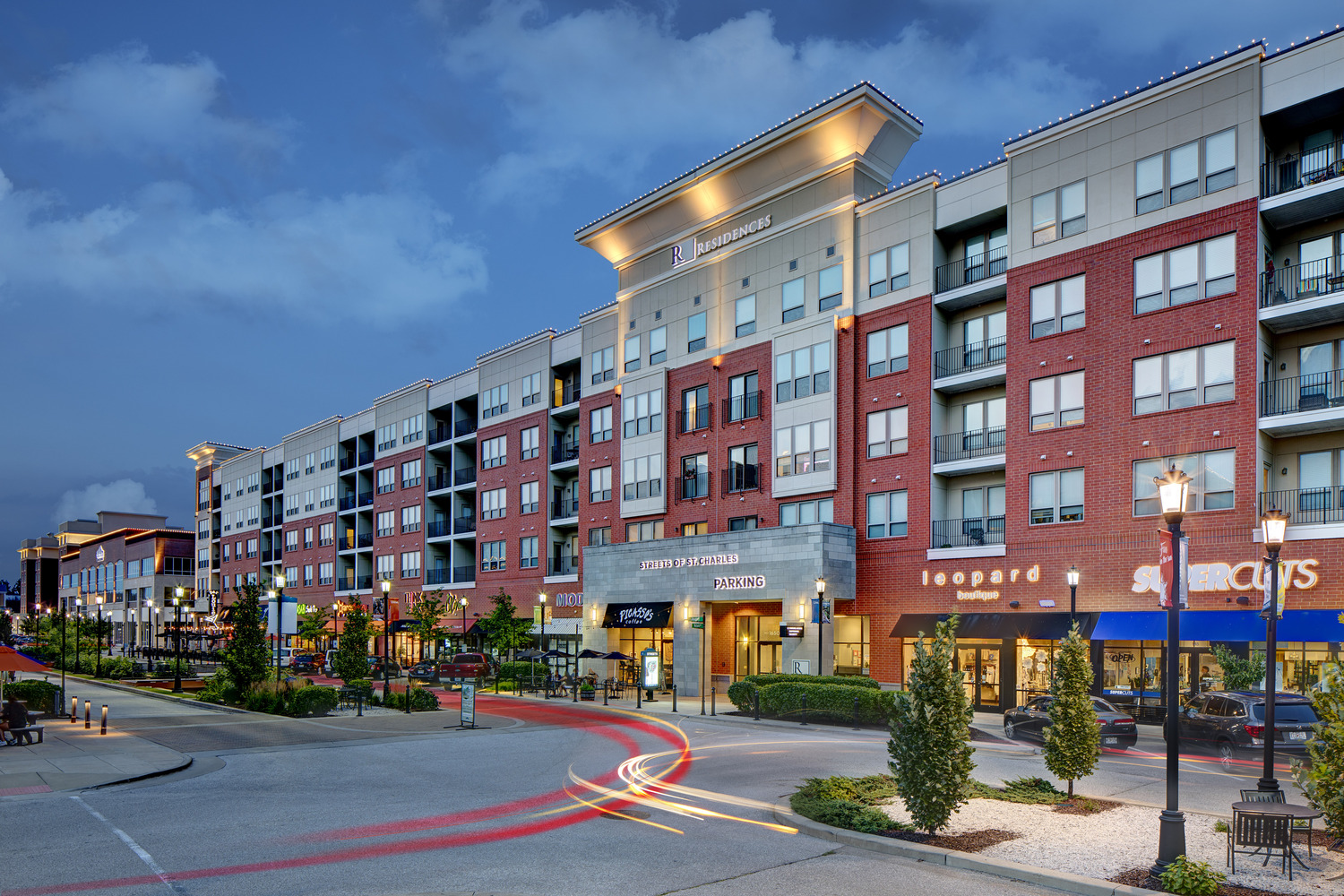 Modern mixed-use building with retail stores and restaurants on the ground floor and multiple stories of apartments above, photographed at dusk with streetlights, trees, and light trails from passing cars in front.