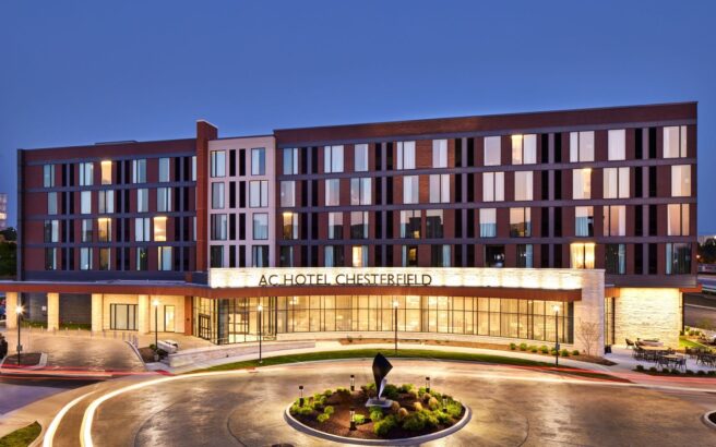 Modern four-story hotel with large windows and illuminated sign reading “AC Hotel Chesterfield” at dusk, featuring a circular driveway with landscaped centerpiece in front.