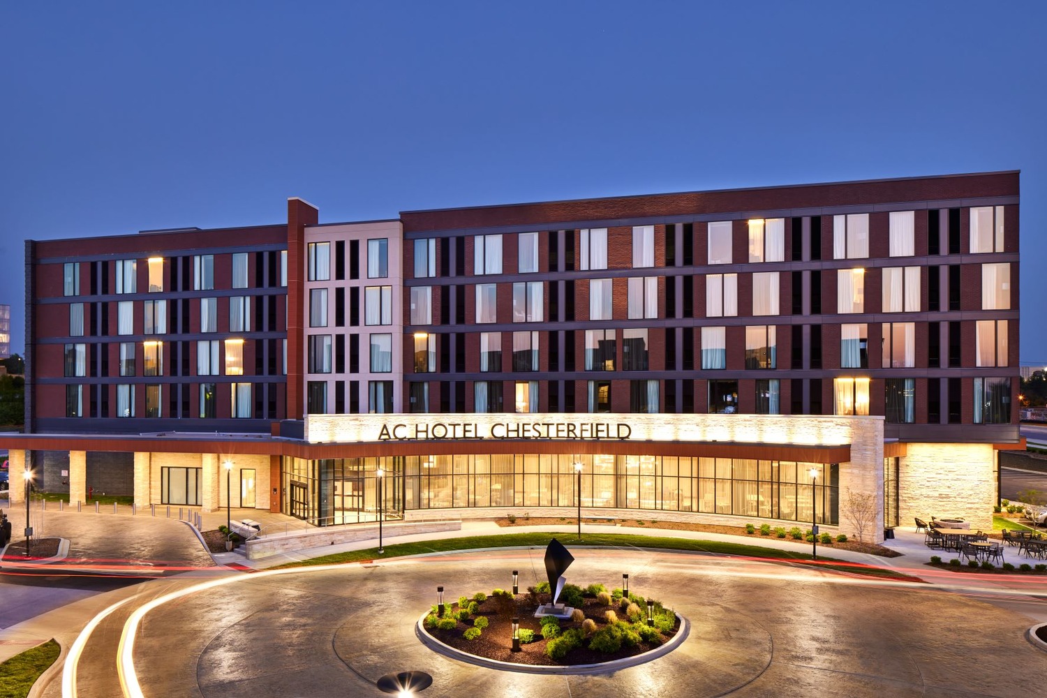 Modern four-story hotel with large windows and illuminated sign reading “AC Hotel Chesterfield” at dusk, featuring a circular driveway with landscaped centerpiece in front.