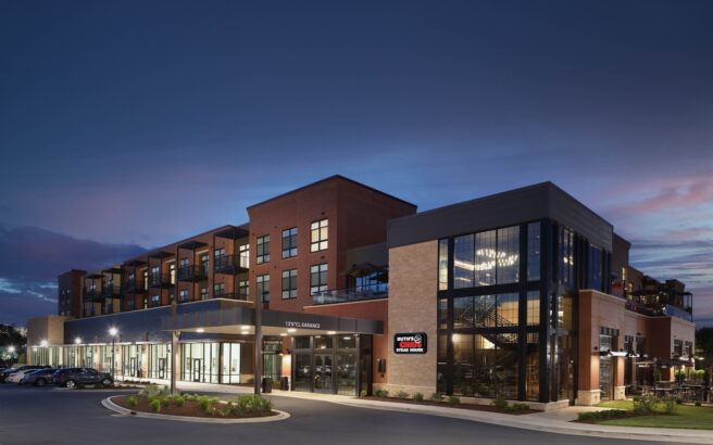 A modern, multi-story building with large windows and balconies, featuring a restaurant on the ground floor, is illuminated at dusk against a deep blue sky. The parking lot and landscaped areas are visible in front.