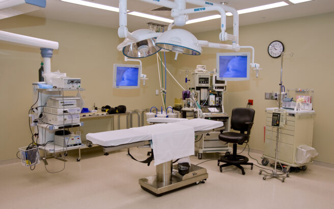 A modern operating room with an empty surgical table, medical equipment, monitors displaying images, overhead surgical lights, a black chair, and various machines against beige walls. The room is clean and brightly lit.