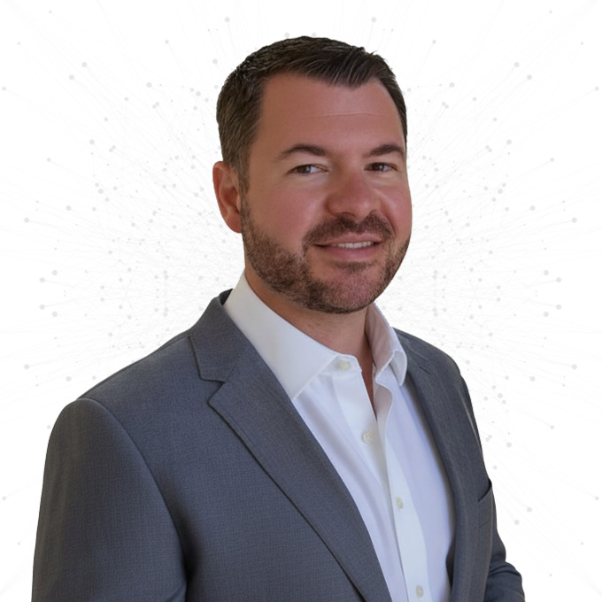 A man with short dark hair and a trimmed beard, wearing a gray suit jacket and white dress shirt, smiles at the camera against a white background with a subtle dotted pattern.