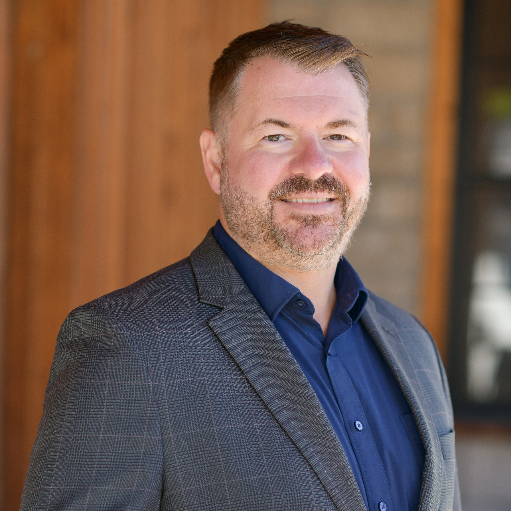 A man with short light brown hair and a beard, wearing a gray checkered blazer over a navy blue shirt, stands smiling in front of a blurred wooden and brick background.