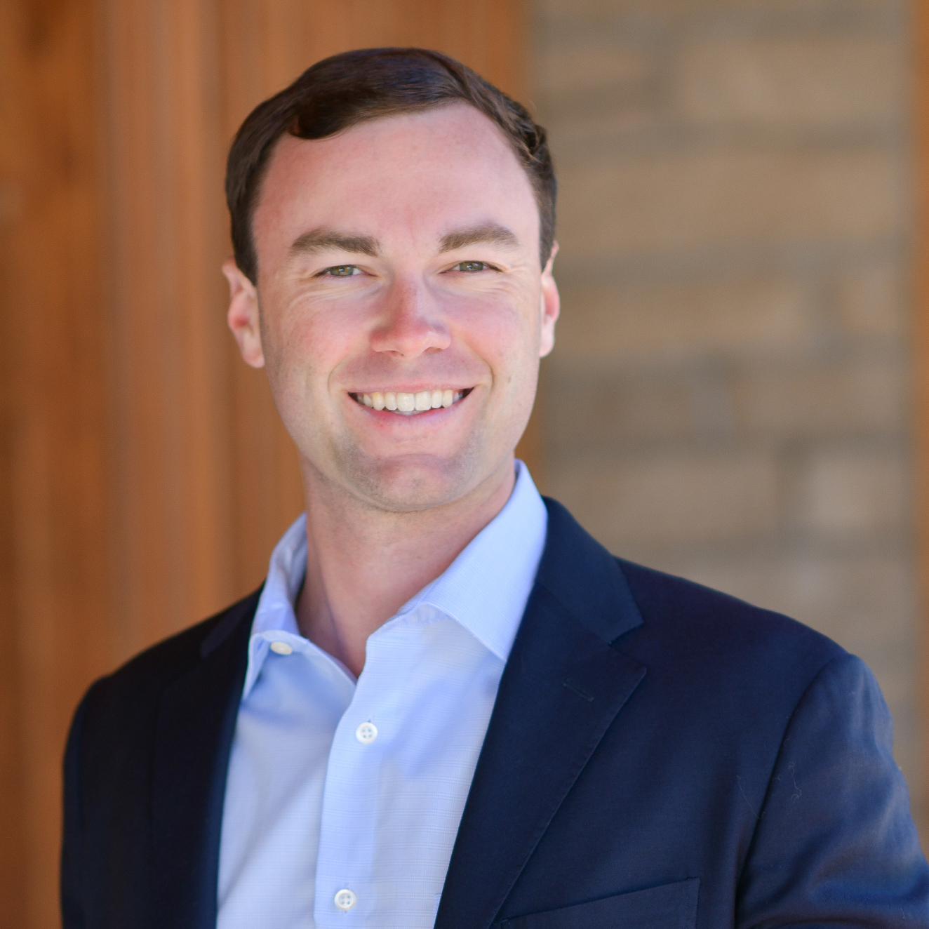 A man with short brown hair wearing a blue dress shirt and dark blazer smiles in front of a blurred background of wooden panels and stone.