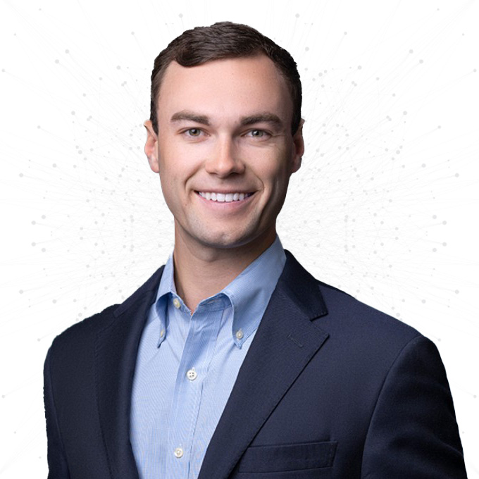 A young man wearing a blue dress shirt and dark blazer smiles at the camera against a white background with subtle abstract dot patterns.