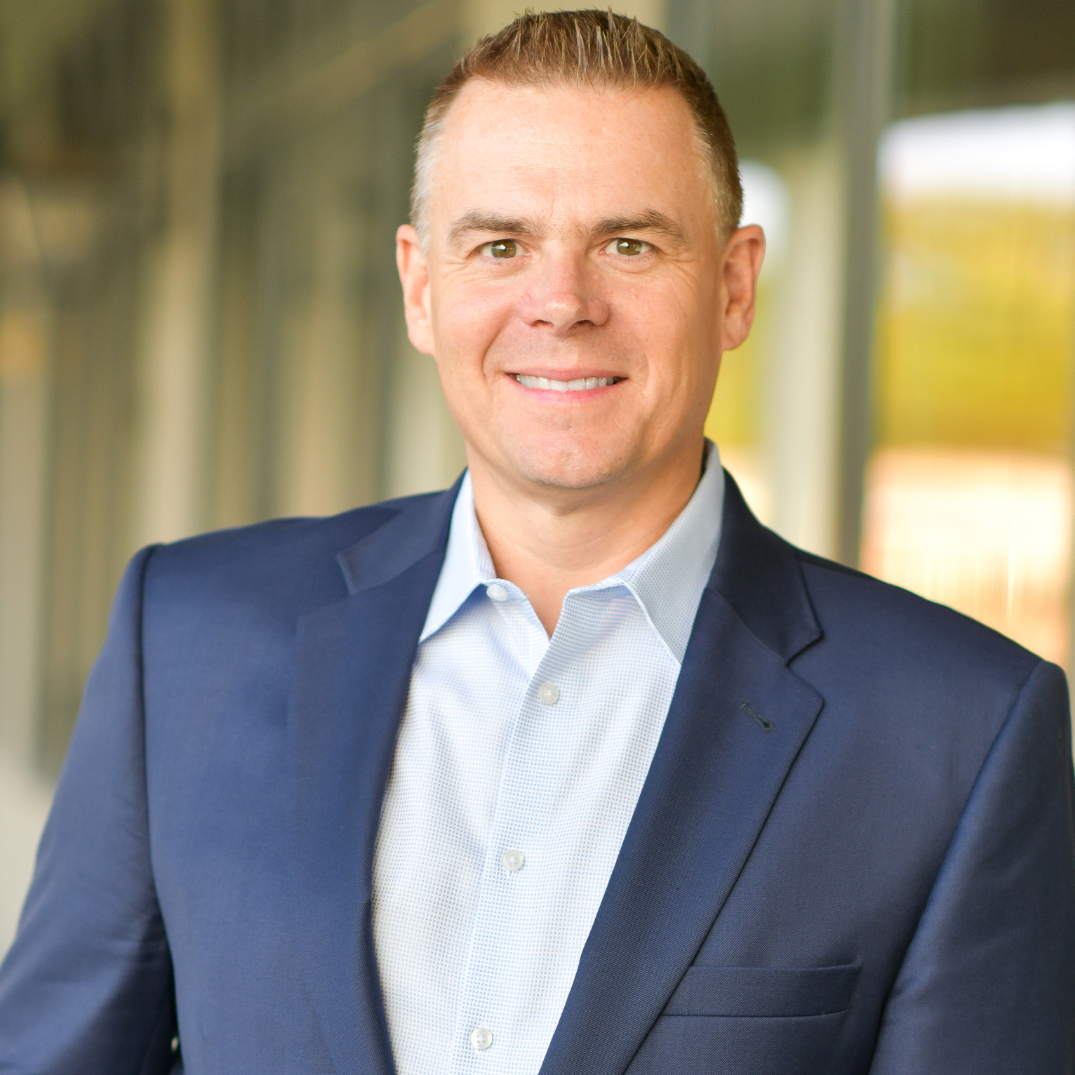 A man in a blue suit jacket and light-colored shirt smiles at the camera while standing in front of a blurred glass building background.