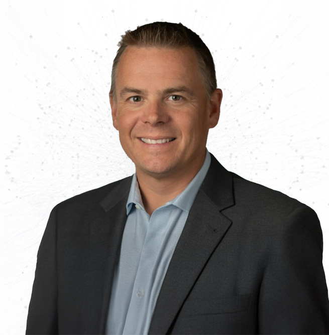 A man with short light brown hair wearing a dark suit jacket over a light blue collared shirt smiles at the camera against a white background with subtle abstract patterns.