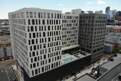 A modern, multi-story apartment building with a central courtyard featuring a swimming pool, surrounded by city streets and other urban buildings under a clear sky.