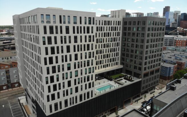 A modern, multi-story apartment building with a central courtyard featuring a swimming pool, surrounded by city streets and other urban buildings under a clear sky.