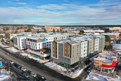 Aerial view of a snowy urban area with modern multi-story apartment buildings, a Chick-fil-A restaurant in the foreground, and other commercial and institutional buildings in the background under a blue sky.
