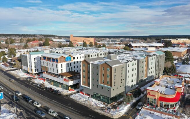Aerial view of a snowy urban area with modern multi-story apartment buildings, a Chick-fil-A restaurant in the foreground, and other commercial and institutional buildings in the background under a blue sky.