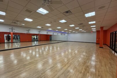 Spacious, empty dance studio with a polished wooden floor, mirrors along one wall, bright ceiling lights, and red and white walls. The room has large windows and a clean, open appearance.
