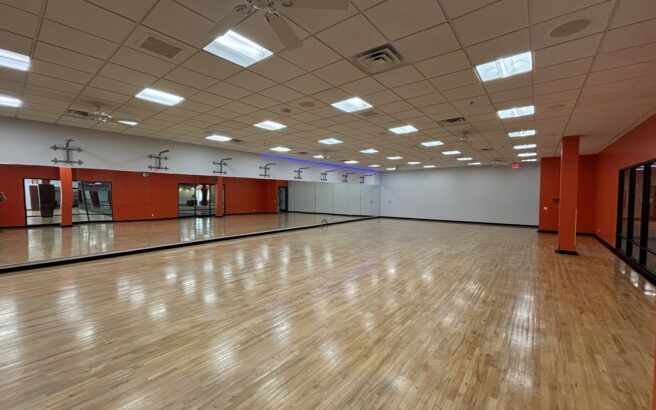Spacious, empty dance studio with a polished wooden floor, mirrors along one wall, bright ceiling lights, and red and white walls. The room has large windows and a clean, open appearance.