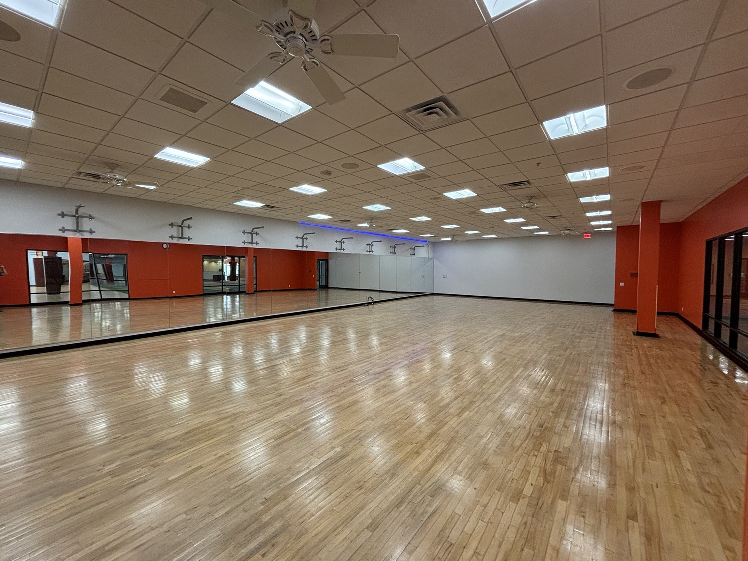 Spacious, empty dance studio with a polished wooden floor, mirrors along one wall, bright ceiling lights, and red and white walls. The room has large windows and a clean, open appearance.