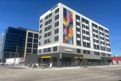A modern six-story hotel building with a colorful geometric mural on one side, large windows, and the word CAMBRiA on the exterior, located on a city street under a clear blue sky.