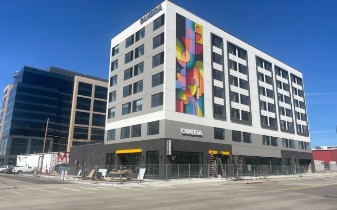 A modern six-story hotel building with a colorful geometric mural on one side, large windows, and the word CAMBRiA on the exterior, located on a city street under a clear blue sky.