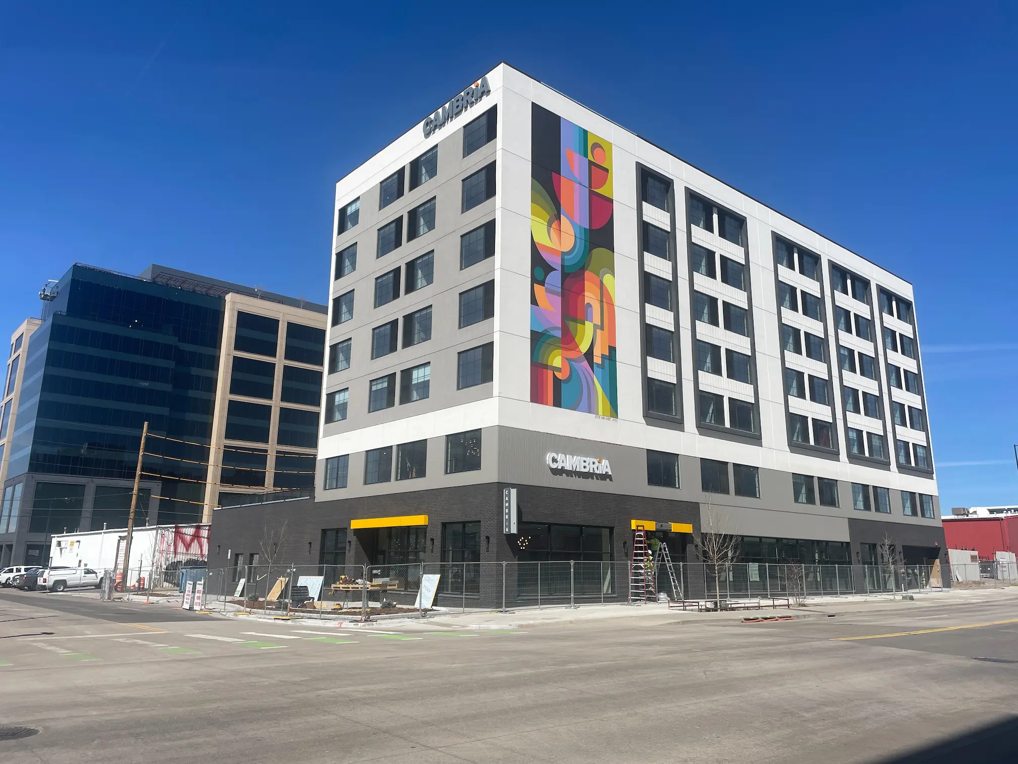 A modern six-story hotel building with a colorful geometric mural on one side, large windows, and the word CAMBRiA on the exterior, located on a city street under a clear blue sky.