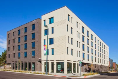 A modern, four-story apartment building with white and dark gray brick facades sits on a city corner under a clear blue sky. Large windows line the exterior, and a street lamp with banners stands near the entrance.