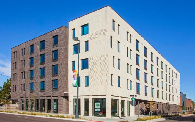 A modern, four-story apartment building with white and dark gray brick facades sits on a city corner under a clear blue sky. Large windows line the exterior, and a street lamp with banners stands near the entrance.