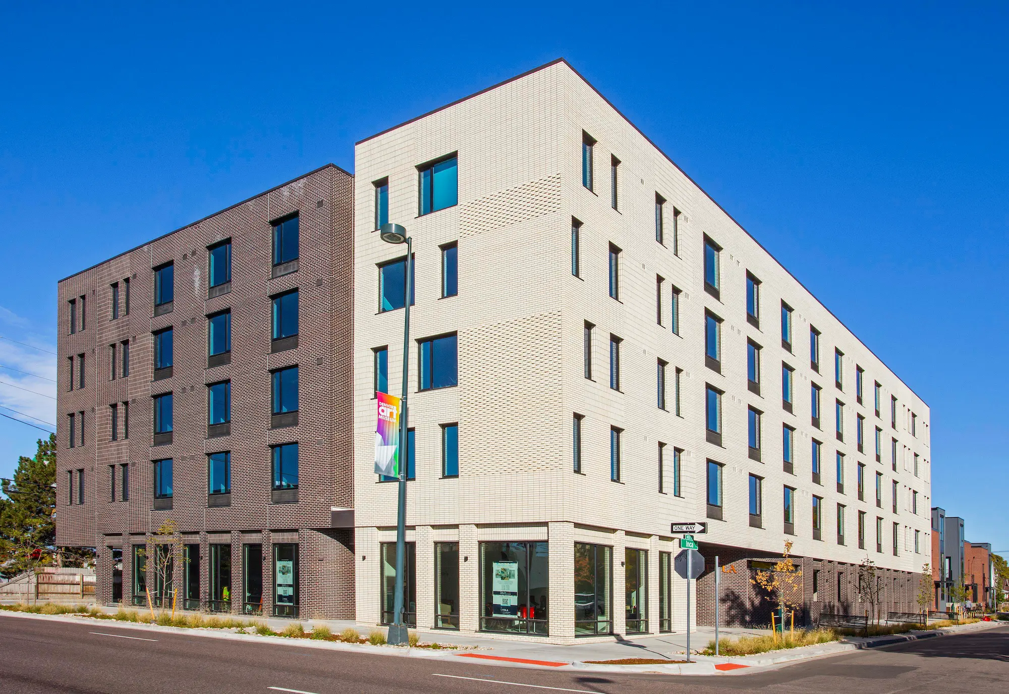 A modern, four-story apartment building with white and dark gray brick facades sits on a city corner under a clear blue sky. Large windows line the exterior, and a street lamp with banners stands near the entrance.