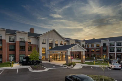 Exterior view of a modern, multi-story residential building with brick and siding at sunset, featuring a covered entrance, parked vehicles, landscaped areas, and illuminated windows.
