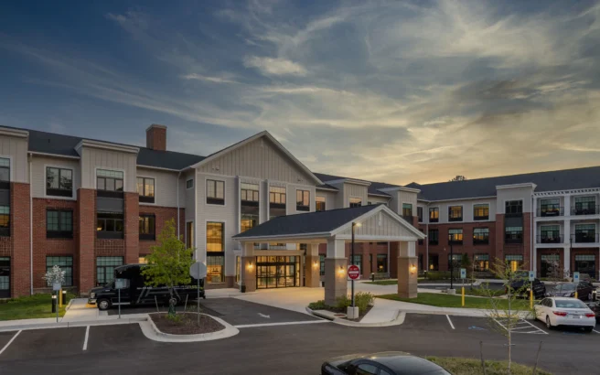Exterior view of a modern, multi-story residential building with brick and siding at sunset, featuring a covered entrance, parked vehicles, landscaped areas, and illuminated windows.